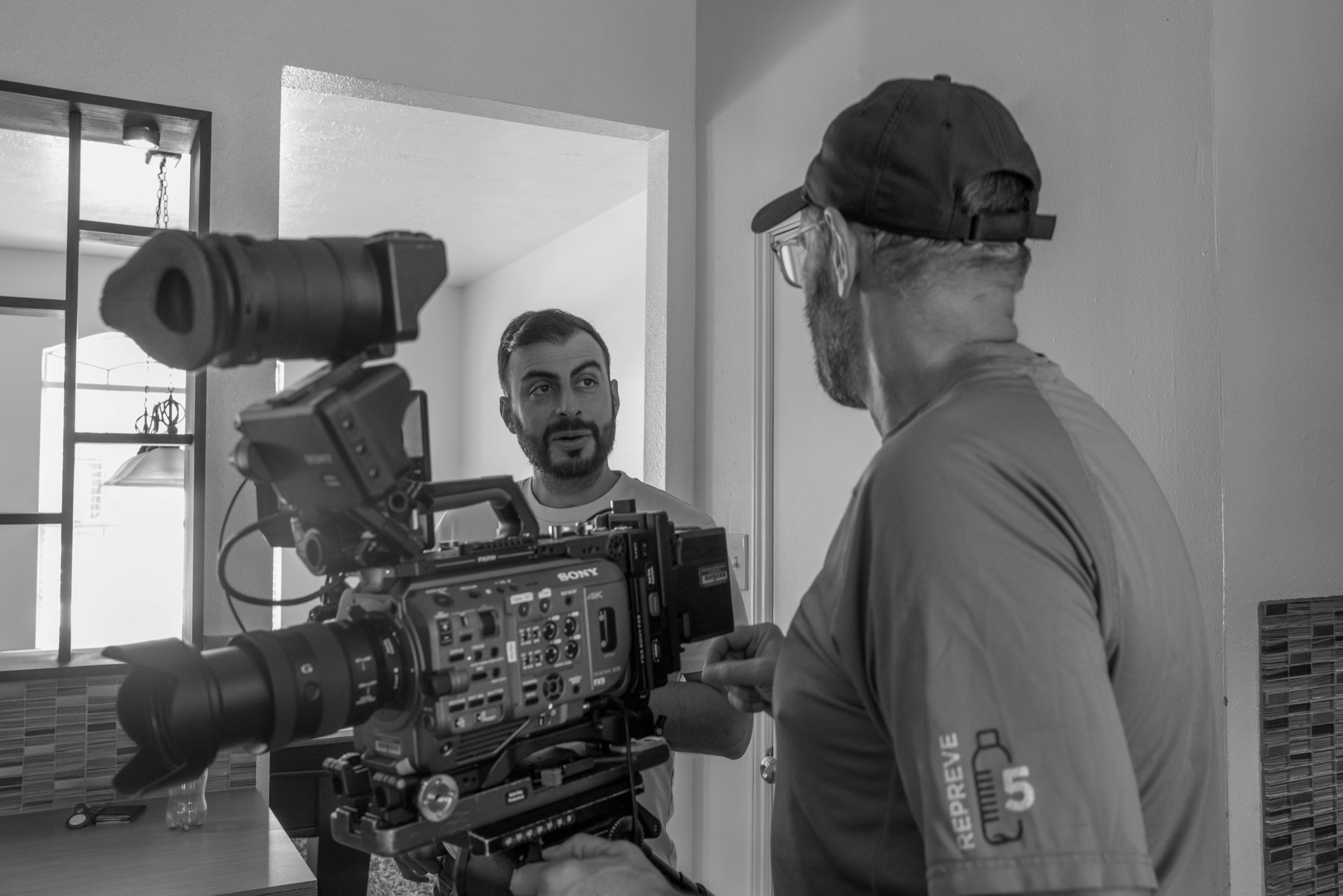 Camera operator framing an interview subject on a Sony cinema camera in a bright kitchen